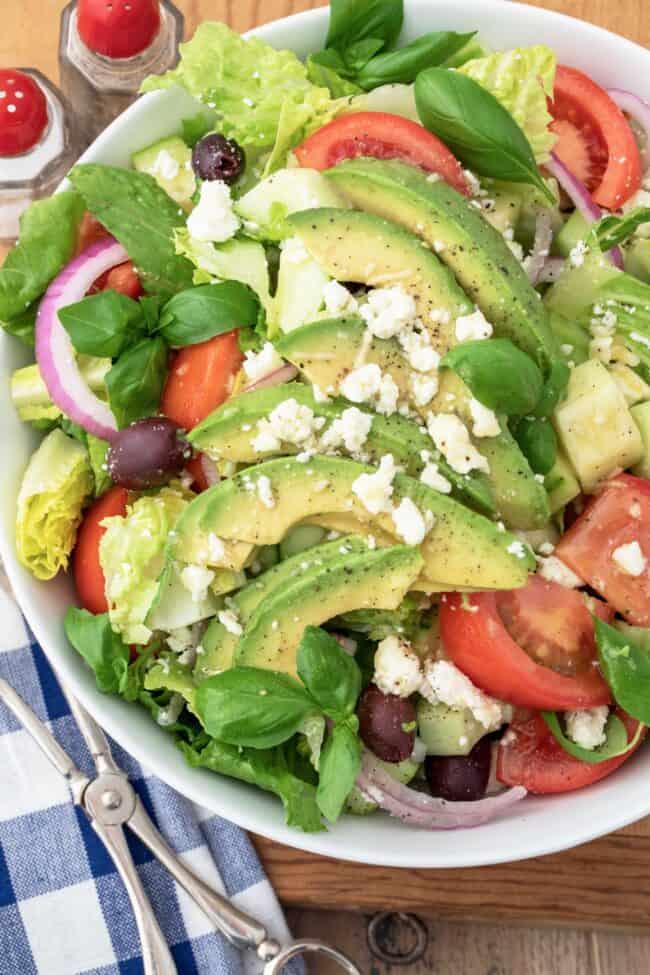 A white bowl of green salad with avocado, tomatoes and kalamata olives